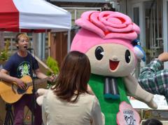 バラの花がモチーフになっているマスコットキャラクターのやっぴーとギターを弾きながら歌う男性の写真