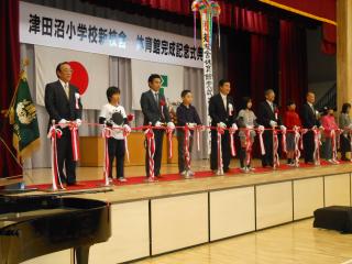 「津田小学校新校舎体育館完成記念式典」と書かれた看板と、舞台で関係者と小学生がテープカットをする写真