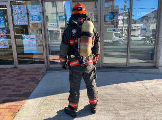 オレンジ色のヘルメットを被り防火衣を着て、空気呼吸器を背中に背負った消防士を真後ろから写した写真