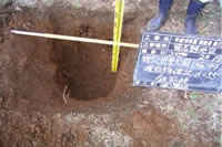 地面が掘られ、目盛りで深さをはかり、穴の横に置かれた黒板と磁気探査で確認された鉄筋棒を写した写真