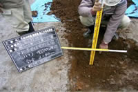地面が掘られ、黒板の横で、深さを測っている作業員と磁気探査で確認された針金の位置を示している写真