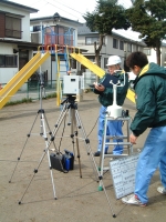 公園内で、2名の職員の方が三脚に機械を設置して環境大気の調査をしている様子の写真