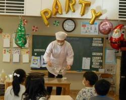 前方の黒板の周りに「PARTY」や「サンタクロース」のバルーンが飾られており、白色の調理衣を着た男性がテーブルの上で作業をしている様子を子供たちが見ている写真