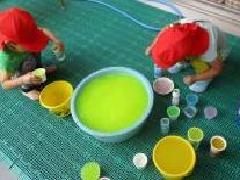 たらいに入っている色水をコップなどに入れてジュース屋さんごっこ遊びをしている2名の園児の写真