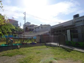 1階建ての建物の軒から所庭にある棒のような物に日陰ネットが設置されている2歳児所庭の写真