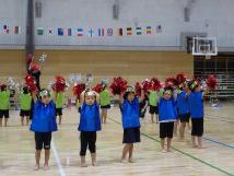青や緑色の服を着た子供たちが赤いポンポンを手に持ち、上に挙げて踊っている様子の写真