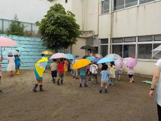 雨の中、傘をさして幼稚園の校庭に集まっている子供たちの写真