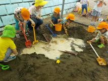 砂場に出来た穴の中の水をスコップやバケツですくって遊んでいる子供たちの写真