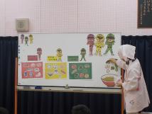 ホワイトボードにラーメンを作る過程を赤、黄色、緑の三色で表現して説明をしている割烹着を着た栄養士の先生の写真