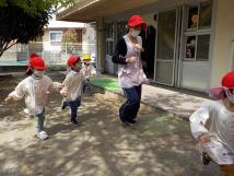 赤い帽子を被った子供たちが先生と一緒に園庭を走っている様子の写真