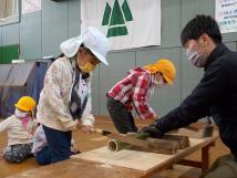 作業机の上でのこぎりを使って竹を切っている子供と作業をサポートする鹿野山の先生の写真