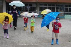 色とりどりの傘をさして園庭で傘の差し方を練習している園児と先生の写真