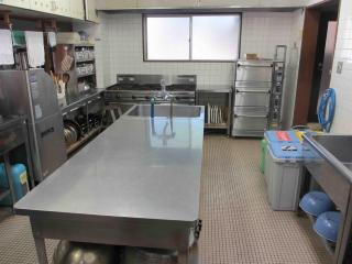 中央に流しの付いた大きな調理台、壁側に冷蔵庫やコンロなどの調理器具が設置されている本館厨房の写真