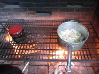 木が重ねて置かれた薪に火が付き、網の上に置かれた小さな丸い缶と深鍋で調理している写真